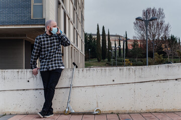 Wall Mural - Hombre joven sentado hablando por teléfono mientras lleva una mascarilla desechable quirúrgica.  Hombre con camisa de cuadros con un teléfono móvil al lado de un patinete no eléctrico de metal.