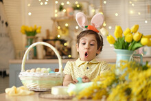 Child Boy Wearing Bunny Ears Sitting In The Kitchen At The Table. Child Holding Painted Eggs Preparing For Easter. Funny Surprised Child