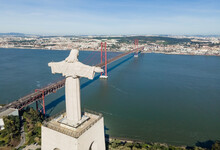 Aerial View Of Christ The King (Cristo Rei) Landmark Statue Facing April 25th Bridge Crossing Tagus River With Lisbon Downtown In Background, Portugal.