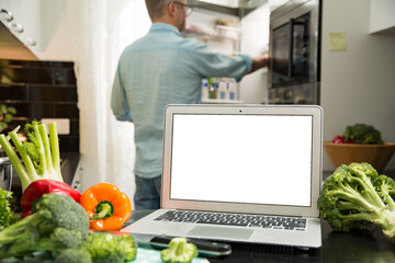 Wall Mural - Man using laptop and cooking in kitchen at home. Distant work. Fresh vegetables on kitchen counter. Healthy lifestyle and social media concept. White empty screen