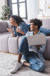 © LIGHTFIELD STUDIOS - Young african american man with laptop looking at cheerful girlfriend with smartphone on couch