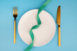 white round plate and golden fork and knife with green measuring tape on blue background, Concept of anorexia and diet