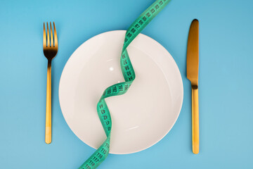 white round plate and golden fork and knife with green measuring tape on blue background, Concept of anorexia and diet