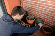 © Edgar - Hombre joven caucásico concentrado sentado en el suelo de su terraza cuidando las plantes que tiene en las macetas. Añadiendo tierra para las plantas