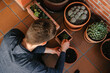 © Edgar - Hombre joven caucásico concentrado sentado en el suelo de su terraza cuidando las plantes que tiene en las macetas. Añadiendo tierra para las plantas