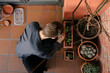 © Edgar - Hombre joven caucásico concentrado sentado en el suelo de su terraza cuidando las plantes que tiene en las macetas. Añadiendo tierra para las plantas