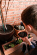 © Edgar - Hombre joven caucásico concentrado sentado en el suelo de su terraza cuidando las plantes que tiene en las macetas. Añadiendo tierra para las plantas