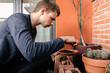 © Edgar - Hombre joven caucásico concentrado sentado en el suelo de su terraza cuidando las plantes que tiene en las macetas. Añadiendo tierra para las plantas