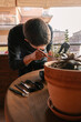 © Edgar - Hombre caucásico joven en la terraza de su casa cuidando sus plantas y suculentas con varias plantas encima de la mesa y material para cuidarlas
