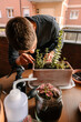 © Edgar - Hombre caucásico joven en la terraza de su casa cuidando sus plantas y suculentas con varias plantas encima de la mesa y material para cuidarlas
