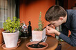 © Edgar - Hombre caucásico joven en la terraza de su casa cuidando sus plantas y suculentas con varias plantas encima de la mesa y material para cuidarlas