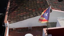 Catalan Flag Flies In The Wind On Sunny Day At The Background Of A Tiled Roof. Slow Motion.