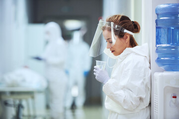 Young surgeon in protective wear sitting in the corridor at hospital and drinking water she resting after operation