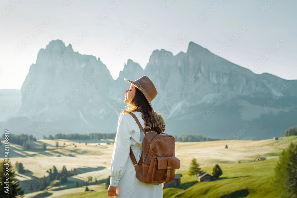 Happy young girl with white dress, hat and backpack in Alpe di Siusi, Dolomites. - obrazy, fototapety, plakaty 