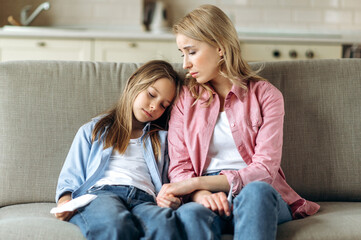 the woman and little girl are sitting at home on the couch. a young blonde mom in sits next to her s