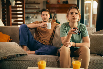 Wall Mural - Young pensive woman watching TV with her boyfriend in the living room.