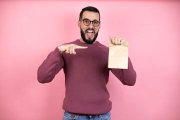 Wall Mural - Handsome man wearing glasses and casual clothes over pink background smiling, surprised and pointing paper bag