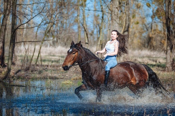 Wall Mural - Beautiful woman rides on the bay the strong horse in the water, raising high sprays. Rider on the horse is moving fast canter on spring forest