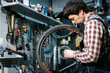 © Elizaveta - Male mechanic working in bicycle repair shop, mechanic repairing bike using special tool, wearing protective gloves. Young attractive serviceman fixing customer's bicycle wheel at his own workshop