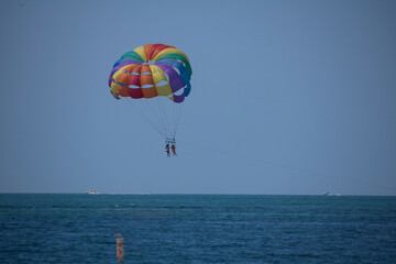 Two people parasailing above blue ocean waters with a boat on the horizon on a sunny summer day with blue skies above.