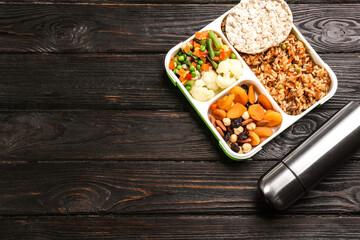 Sticker - Thermos bottle and lunch box with food on black wooden background, flat lay. Space for text