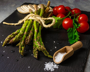 Wall Mural - Composition of various vegetables with bunch of asparagus, mushrooms, cherry tomatoes and parsley, with wooden spoon containing salt on slate surface