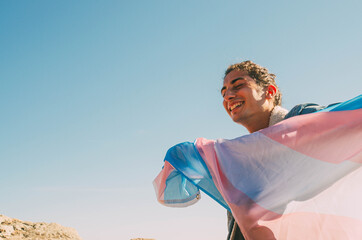 Happy Young Man Smiling While Holding Transgender LGTB Flag With Open Arms and Blue Sky Background