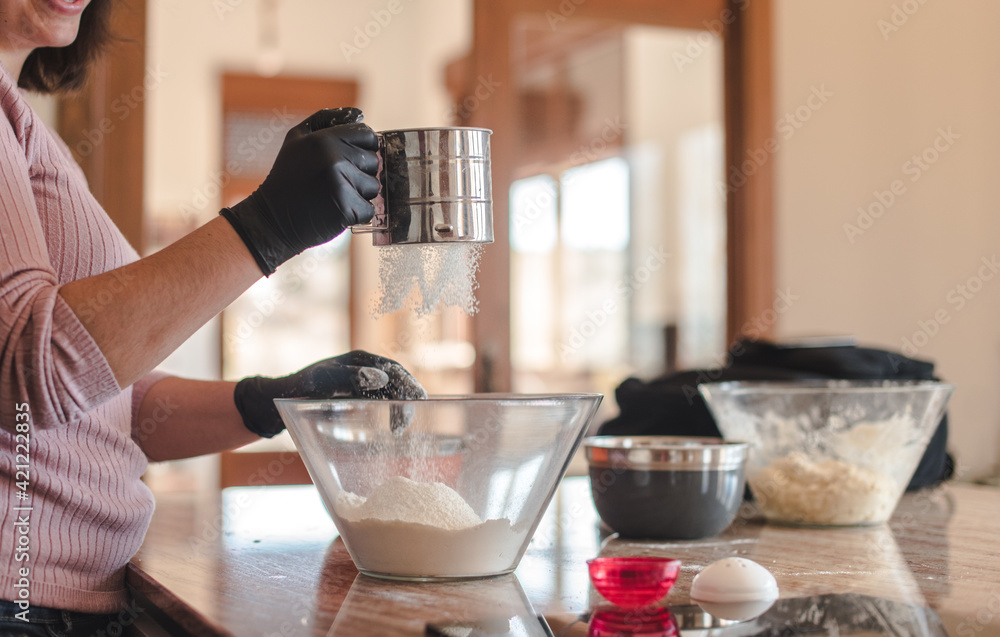 Mujer joven con guantes en su cocina mientras tamiza la harina en un recipiente de cristal. Concepto de cocinar pizza casera. Hacer pan en casa. - obrazy, fototapety, plakaty 