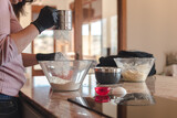 Fototapeta  - Mujer joven con guantes en su cocina mientras tamiza la harina en un recipiente de cristal. Concepto de cocinar pizza casera. Hacer pan en casa.