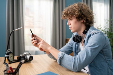 Wall Mural - Portrait of a Caucasian Teenage Boy with Curly Hair in Cozy Living Room Using Smartphone at Home. He is Browsing the Internet and Checking Videos on Social Networks and Typing message.