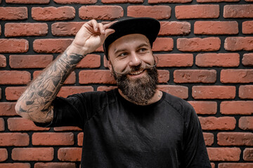 Handsome old-fashioned hipster in t-shirt and cap, looks happy and smile. On a brick background.