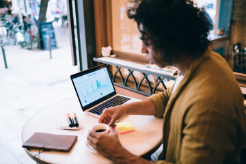 Wall Mural - Serious businessman in casual wear using laptop computer for checking revenue and income via infographic on monitor, pensive smart 20s male student learning homework in cafeteria via devices