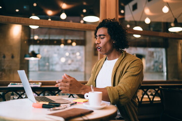 Wall Mural - Handsome emotional 20s hipster guy watching videos on laptop computer sitting in cafe interior,expressive young millennial man surprised with news from social networks pointing on monitor