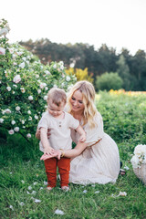 Young beautiful blond mother in white dress and toddler son outdoor portrait in summer park. Flower bushes of roses. Nature, warm weather, summer fashion clothes.