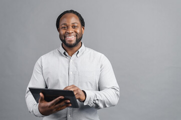 Cheerful African-American young man with dreadlocks holding a digital tablet, testing computer app, is surfing in networks, scrolling news feed in social networks, isolated on grey background