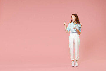 Full length side view young surprised redhead woman in blue shirt pants using mobile cell phone point index finger aside on workspace area mock up isolated on pastel pink background studio portrait