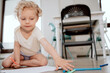 © ABCreative - A little girl paints with markers while sitting on the wooden floor in the living room of an apartment.