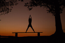 Silhouette Woman With Arms Raised Exercising On Bench Against Sky During Sunset