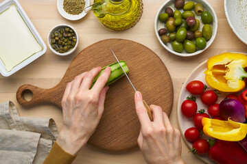 Wall Mural - Vegetarian food. Chopping cucumber, cutting vegetables for greek salad horiatiki. Woman hands with knife on wooden cutting board slicing chop of vegetables. Homemade vegan food, top view