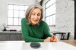 © Vadim Pastuh - Mature pretty woman with gray hair in green jumper talking to the digital virtual assistant at home, asking a question or requesting to switch music. Smart AI speaker concept and voice command control