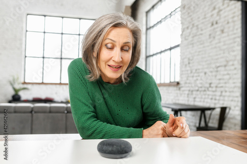 Mature pretty woman with gray hair in green jumper talking to the digital virtual assistant at home, asking a question or requesting to switch music. Smart AI speaker concept and voice command control