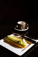 Wall Mural - A vertical photo of a pistachio eclair on a white plate, dessert knife and fork and a cup of coffee in the bokeh. Dark table.