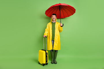 Poster - Full size photo of brunet optimistic guy with umbrella bag wear cloak boots isolated on green color background