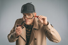 Male Detective With Magnifying Glass Smoking Pipe On Grey Background
