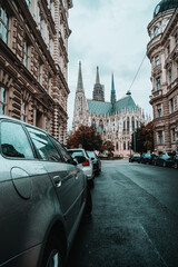 Beautiful streets of Austrian capital Vienna. Old gothic architecture around the narrow streets. Wet asphalt after rain and cars parked around. View on Catholic cathedral Votivkirche in downtown.