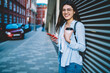 © BullRun - Half length portrait of cheerful woman with coffee to go holding mobile phone smiling at camera at urban settings, funny Caucasian female posing at city street using cellphone technology during travel