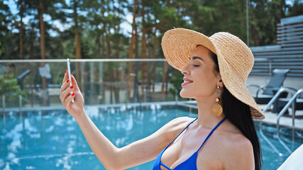 Wall Mural - happy brunette woman in straw hat taking selfie near pool