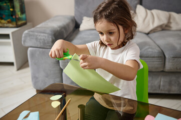 Adorable little preschool girl with braids sitting at the table and concentrated on cutting shapes from color green paper.