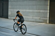 Rear view of professional female cyclist in black cycling garment and protective gear riding bicycle in city, passing buildings while training outdoors on a daytime