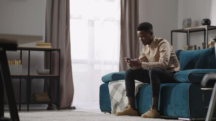 afro-american man is using smartphone for communication and playing games, sitting at home alone, bl